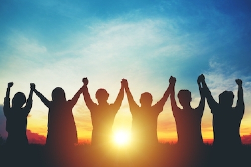 Silhouettes of a diverse group of people standing in a row with hands raised and joined, celebrating together at sunset.